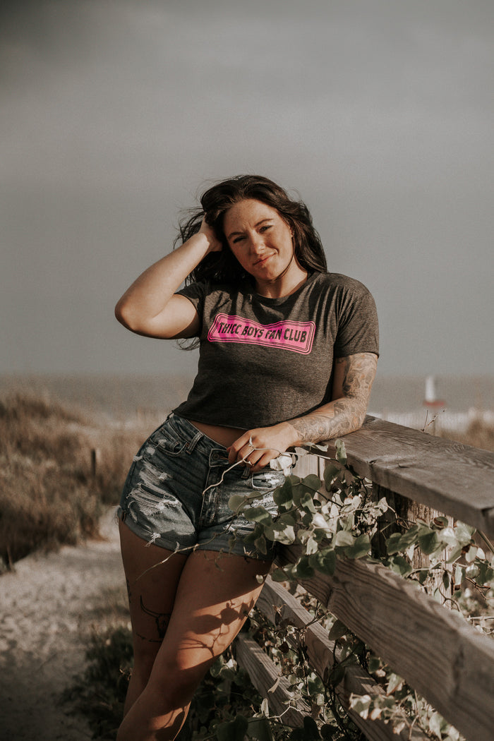 A woman with long dark hair leans on a wooden beach fence, wearing a charcoal Tango Charlie Apparel THICC Boys Fan Club Crop Tee with a pink logo and denim shorts. She looks at the camera, hand in her hair, sand dunes behind her.