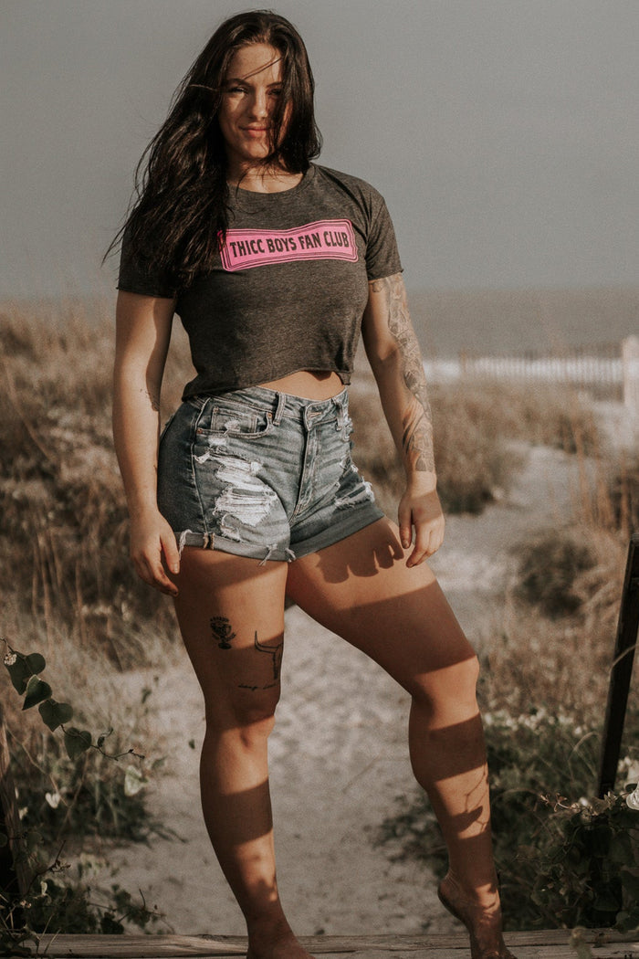 A woman with long dark hair stands barefoot on a sandy beach path, wearing Tango Charlie Apparel's THICC Boys Fan Club Crop Tee in charcoal and distressed denim shorts, sunlight casting shadows on her legs.