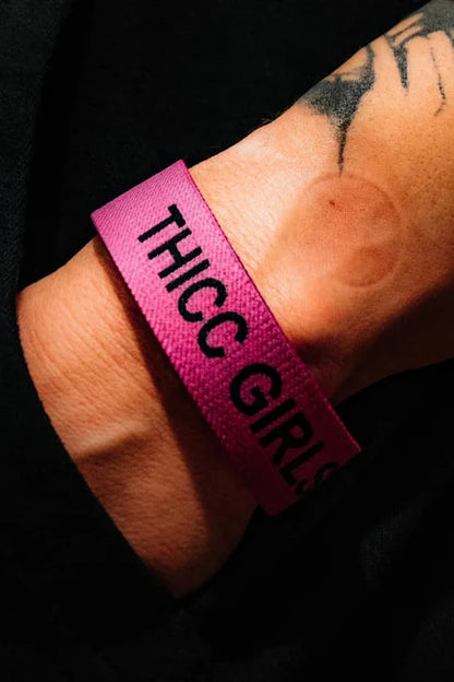 A tattooed wrist showcases the Thicc Girls Club Wristband by Tango Charlie Apparel, featuring bold black THICC GIRLS text on a pink band. Set against a dark backdrop, it celebrates body positivity with every glance.