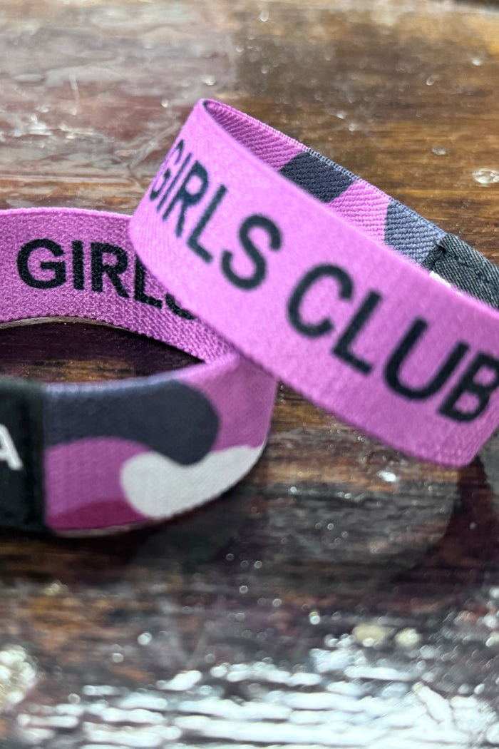 Two purple wristbands from Tango Charlie Apparels Thicc Girls Club rest on wood. One is upright, showing THICC GIRLS CLUB in a black camouflage pattern, symbolizing body positivity. The other lies flat with TCA in white on black—ideal for fans of empowering ladies’ apparel.