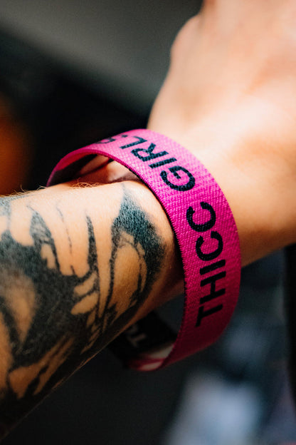 A close-up of a tattooed arm wearing the Thicc Girls Club Wristband by Tango Charlie Apparel—a pink band with "THICC" in bold black letters, perfect for club members.