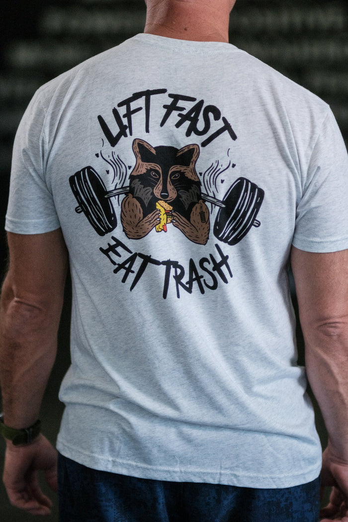 Seen from the rear, a person wears a light gray Trash Panda Weight Lifting Club Tee by Tango Charlie Apparel. It features a trash panda lifting a barbell and holding a banana with LIFT FAST EAT TRASH boldly written. A dark, slightly blurred background adds an edgy vibe.
