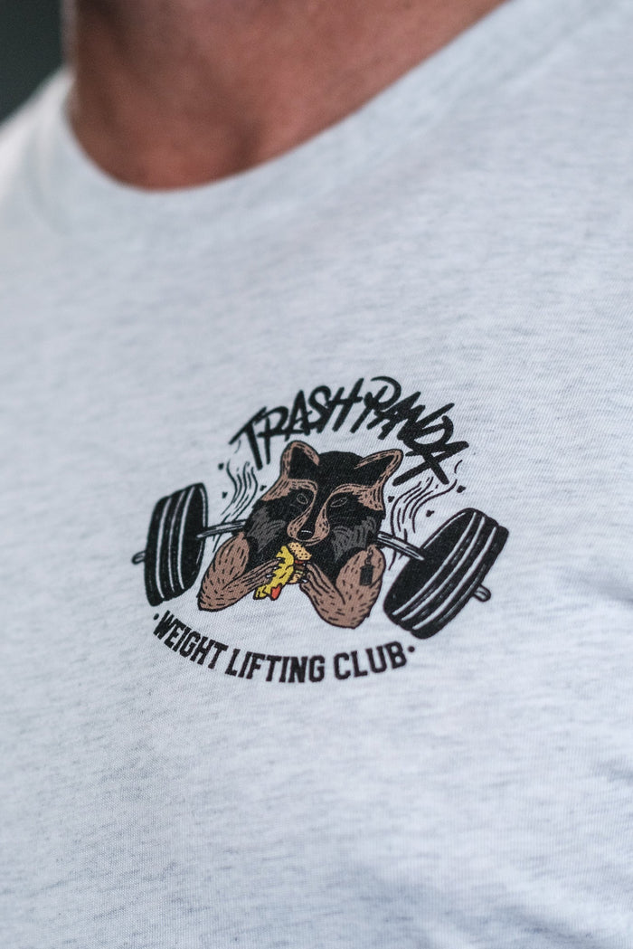A close-up showcases a person in the Tango Charlie Apparels light gray tri-blend Trash Panda Weight Lifting Club Tee featuring a playful raccoon lifting weights logo encircled by the club’s name, combining strength with style.