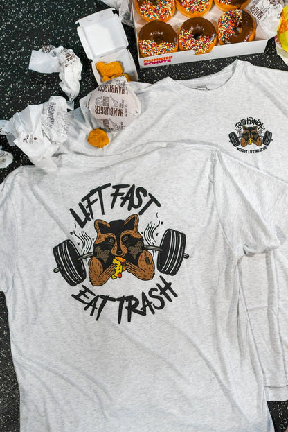 The Trash Panda Weight Lifting Club Tee by Tango Charlie Apparel features a gray tri-blend design with a trash panda lifting weights and eating a burger. Surrounding the Lift Fast, Eat Trash text are fast food items like donuts, wrappers, and nuggets on a speckled surface.