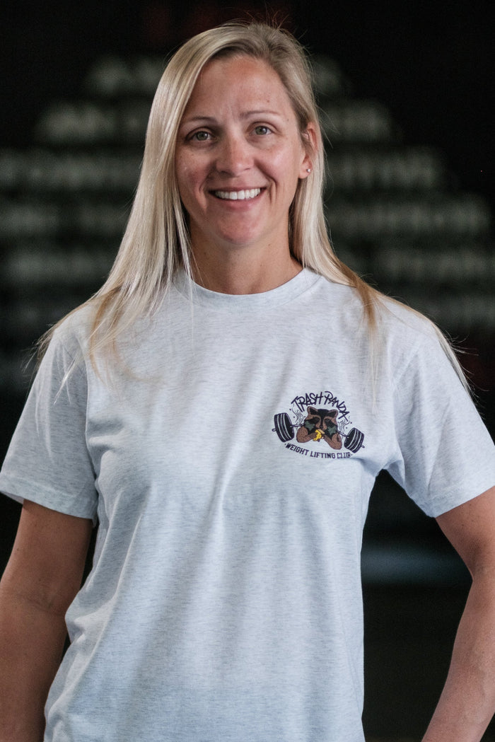 A smiling person with long blonde hair wears a Tango Charlie Apparel Trash Panda Weight Lifting Club Tee, light gray and tri-blend, against a dark, blurred background highlighting a pyramid of weights for an energetic vibe.