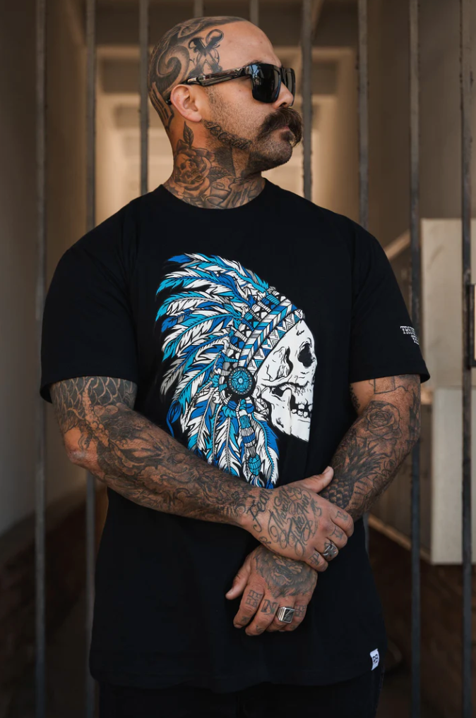 A tattooed man with a mustache and sunglasses, arms crossed, wears the Relentless Betrayal Trustless Chief BLACK T-Shirt featuring a skull in a blue and white feathered headdress. Vertical metal bars form the background.