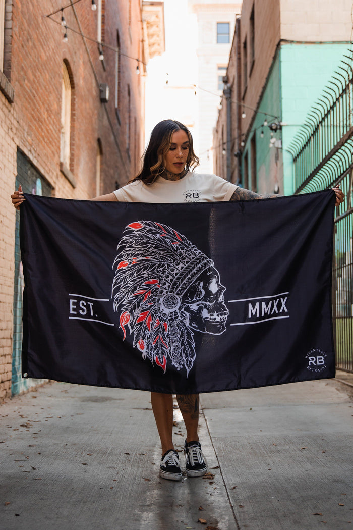 A person stands in an alley holding the Relentless Betrayal Trustless Chief Flag. Its black and red, featuring a skull with a Native American headdress, red and white details, and EST. MMXX. Made of durable polyester with metal grommets, brick walls and iron fencing line the alley.