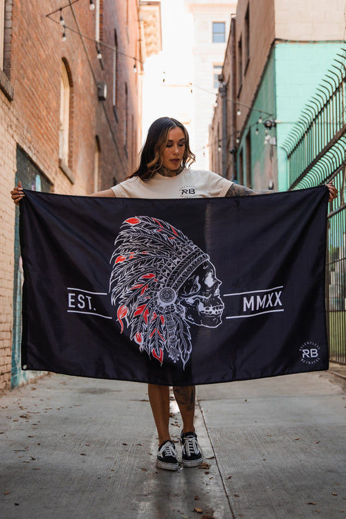 A person stands in an alley holding the Relentless Betrayal Trustless Chief Flag. Its black and red, featuring a skull with a Native American headdress, red and white details, and EST. MMXX. Made of durable polyester with metal grommets, brick walls and iron fencing line the alley.
