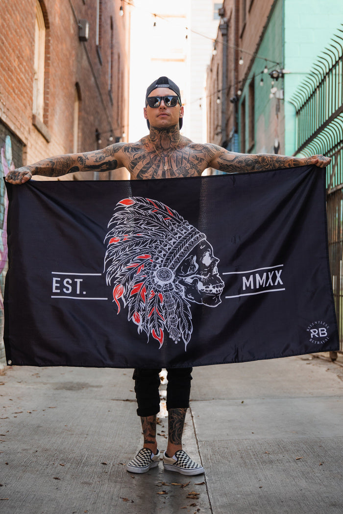 In an urban alley, a tattooed person holds the Trustless Chief Flag by Relentless Betrayal. Made of durable polyester with metal grommets, it features a skull in a feathered headdress and EST. MMXX. They also wear sunglasses, a cap, and checkered shoes against brick walls.