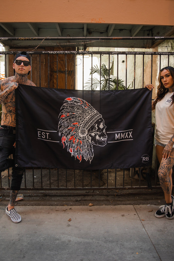 Two tattooed individuals casually dressed hold a Trustless Chief Flag by Relentless Betrayal. The black and red polyester flag features a skull with a Native American headdress adorned in vibrant feathers, metal grommets, and the text Est. MMXX.
