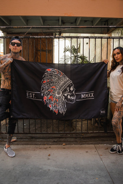 Two tattooed individuals casually dressed hold a Trustless Chief Flag by Relentless Betrayal. The black and red polyester flag features a skull with a Native American headdress adorned in vibrant feathers, metal grommets, and the text Est. MMXX.
