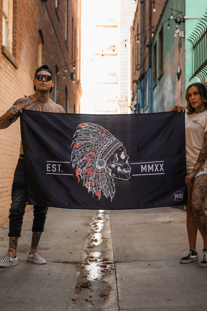 In an alley, two tattooed individuals hold the Relentless Betrayal Trustless Chief Flag, a black and red polyester design showcasing a skull with a feathered headdress and EST. MMXX text. Metal grommets ensure durability as string lights brighten the surrounding brick walls.