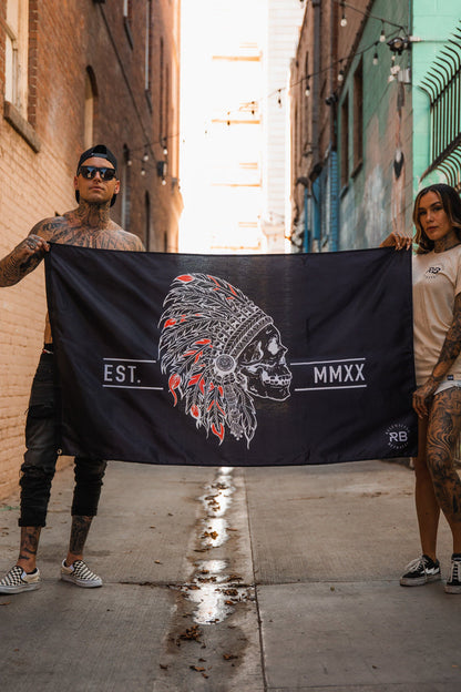 In an alley, two tattooed individuals hold the Relentless Betrayal Trustless Chief Flag, a black and red polyester design showcasing a skull with a feathered headdress and EST. MMXX text. Metal grommets ensure durability as string lights brighten the surrounding brick walls.