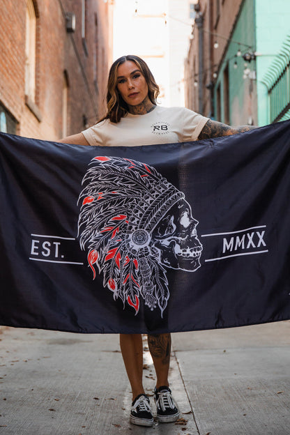 In an alleyway with brick and teal walls, a person in a beige t-shirt and black sneakers holds the Trustless Chief Flag by Relentless Betrayal, featuring a skull in a feathered headdress labeled EST. MMXX. Made of durable polyester with metal grommets for longevity.