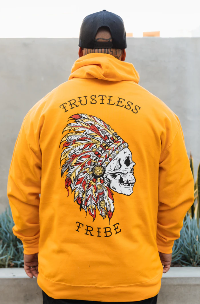 A person wears the Trustless Chief Gold Hoodie, showcasing a vibrant skull with a feather headdress and Trustless Tribe text. Made by Relentless Betrayal, this unisex hoodie combines comfort and style with its soft cotton/polyester blend.