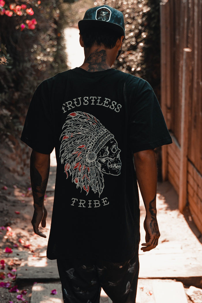 A tattooed person walks outside in a Relentless Betrayal Trustless Chief RED T-Shirt—a unisex black and red fine jersey tee featuring a graphic skull with feathered headdress and TRUSTLESS TRIBE on the back—and wears a black cap.