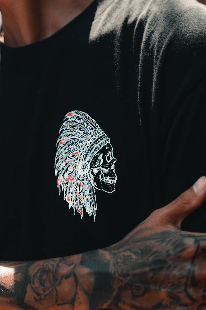 Wearing the Relentless Betrayal Trustless Chief RED T-Shirt—unisex, black and red with a skull in a feathered headdress design—this person shows off a tattooed arm. Made from fine jersey cotton for all-day comfort.