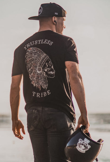 Wearing the Relentless Betrayal Trustless Chief RED T-Shirt, a man stands on the beach with a black helmet, facing away. The unisex tee’s skull and feather headdress graphic adds bold edge in black and red jersey cotton.