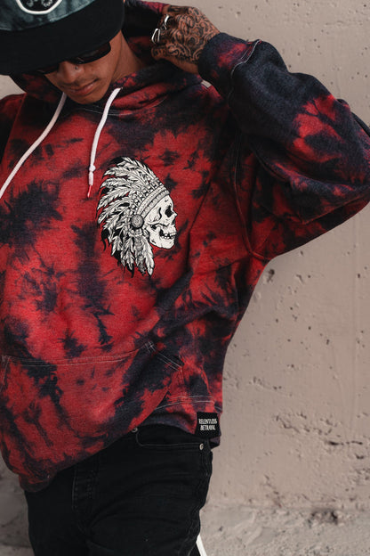 Donning the Trustless Chief Red Tie Dye Hoodie by Relentless Betrayal, featuring a skull and feather headdress design, a person adjusts their hoodie. Tattoos peek out as they sport sunglasses and a cap, blending edgy style with comfort in this unisex cotton/polyester hoodie.