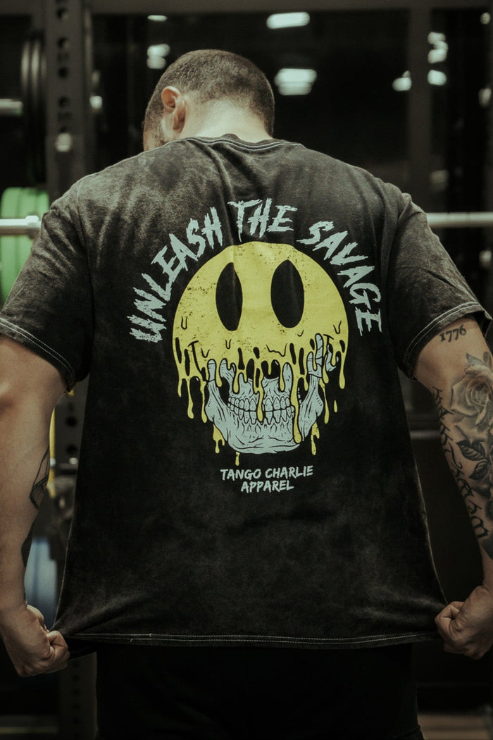 A tattooed person faces away, wearing the Unleash The Savage Premium Tee by Tango Charlie Apparel, which features a large melting yellow smiley over a skull and the phrases "UNLEASH THE SAVAGE" and "TANGO CHARLIE APPAREL" on the back.