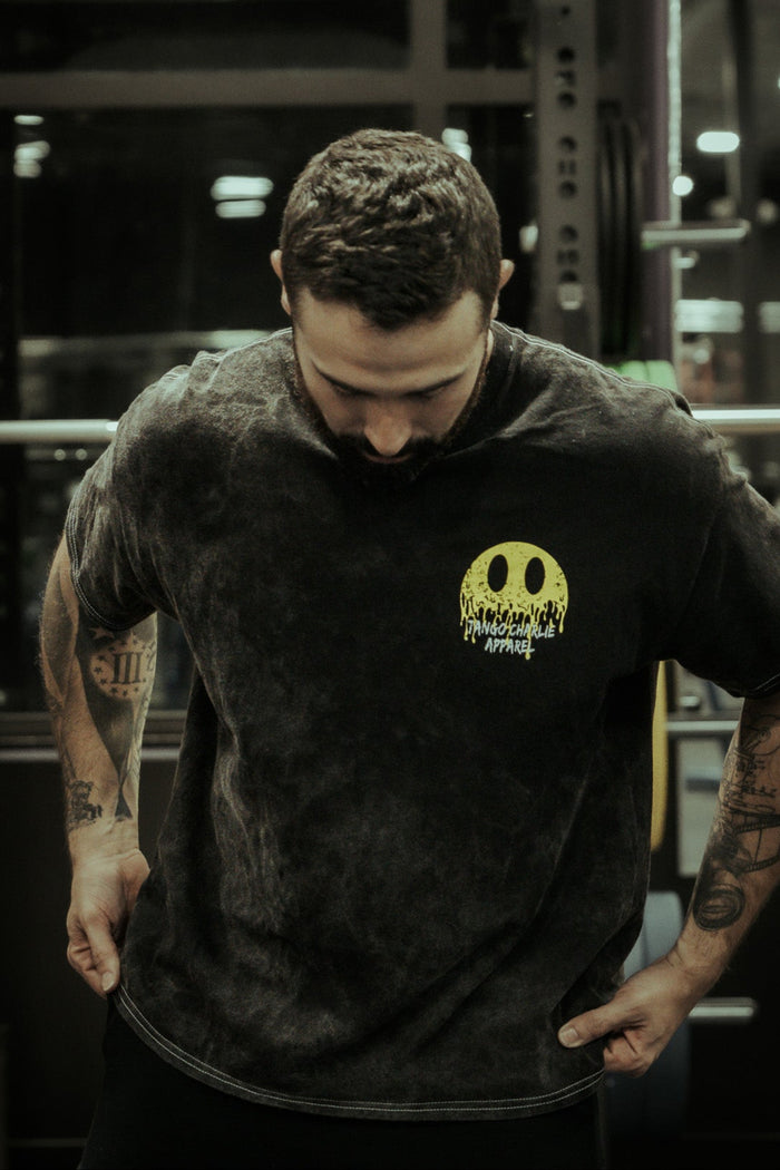 A tattooed man stands in a gym, wearing the Tango Charlie Apparel "Unleash The Savage Premium Tee," featuring a large yellow melting smiley face and text on the chest.