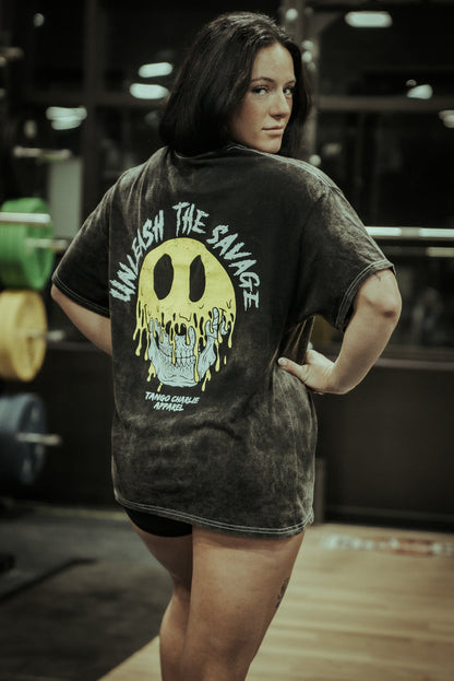 A woman stands in a gym, looking over her shoulder, wearing the Tango Charlie Apparel Unleash The Savage Premium Tee—black with a bold yellow skull and "UNLEASH THE SAVAGE" on the back. Gym equipment fills the background.