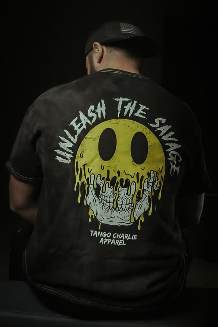 Wearing a dark cap and the Unleash The Savage Premium Tee by Tango Charlie Apparel, featuring a bold yellow melting smiley face and skull graphic. The back displays "UNLEASH THE SAVAGE" and "TANGO CHARLIE APPAREL.