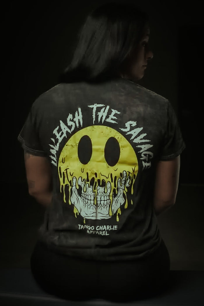 Someone sits facing away, wearing the Unleash The Savage Premium Tee by Tango Charlie Apparel, featuring a melting yellow smiley revealing skulls and edgy text. A dim background highlights the shirt's bold vibe.