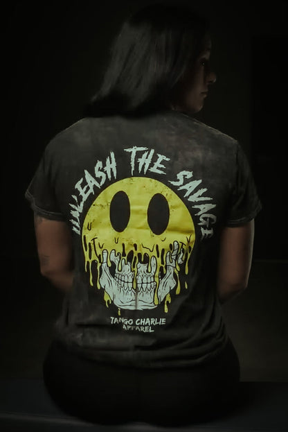 Someone sits facing away, wearing the Unleash The Savage Premium Tee by Tango Charlie Apparel, featuring a melting yellow smiley revealing skulls and edgy text. A dim background highlights the shirt's bold vibe.