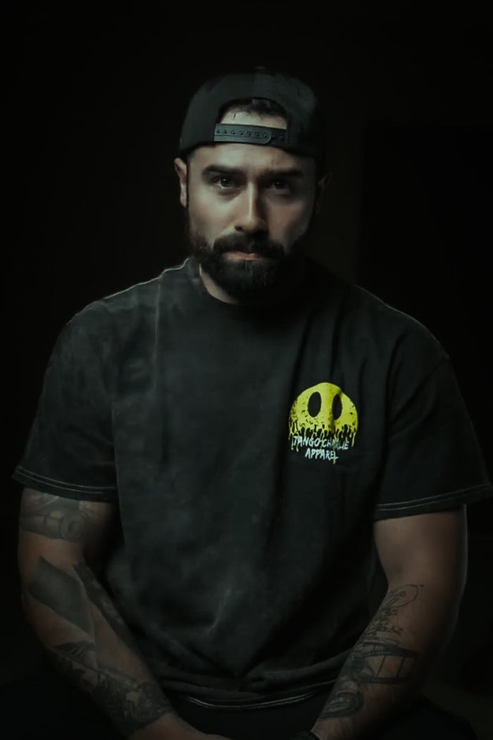 A bearded man with tattooed arms sits facing forward, wearing a Tango Charlie Apparel Unleash The Savage Premium Tee with a bold yellow graphic and a black cap worn backward, his edgy style standing out against the dark background.