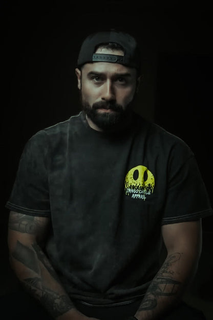 A bearded man with tattooed arms sits facing forward, wearing a Tango Charlie Apparel Unleash The Savage Premium Tee with a bold yellow graphic and a black cap worn backward, his edgy style standing out against the dark background.