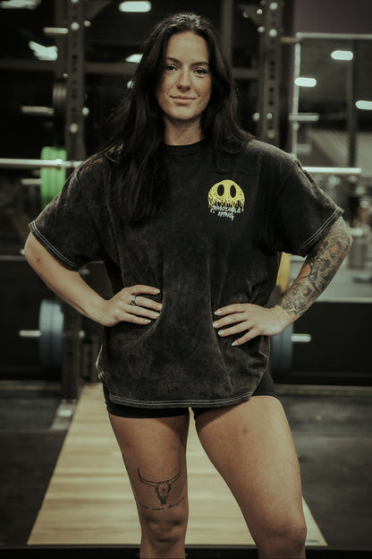 A woman with long dark hair, showing tattoos on her right arm and left thigh, stands confidently in a gym wearing the Tango Charlie Apparel "Unleash The Savage Premium Tee." Weightlifting equipment is visible in the background.