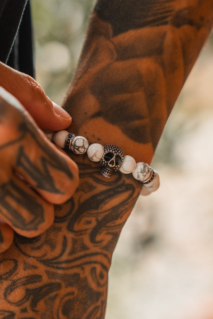 A tattooed hand wears the Relentless Betrayal Until Death Storm Cloud Bead Bracelet, featuring a decorative skull centerpiece and white marbled beads, shown in close-up detail.