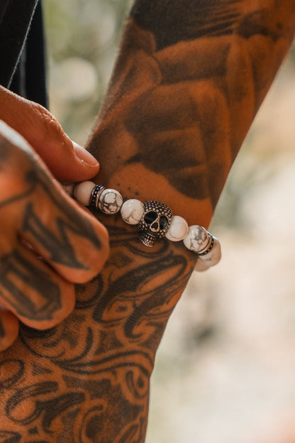 A tattooed hand wears the Relentless Betrayal Until Death Storm Cloud Bead Bracelet, featuring a decorative skull centerpiece and white marbled beads, shown in close-up detail.