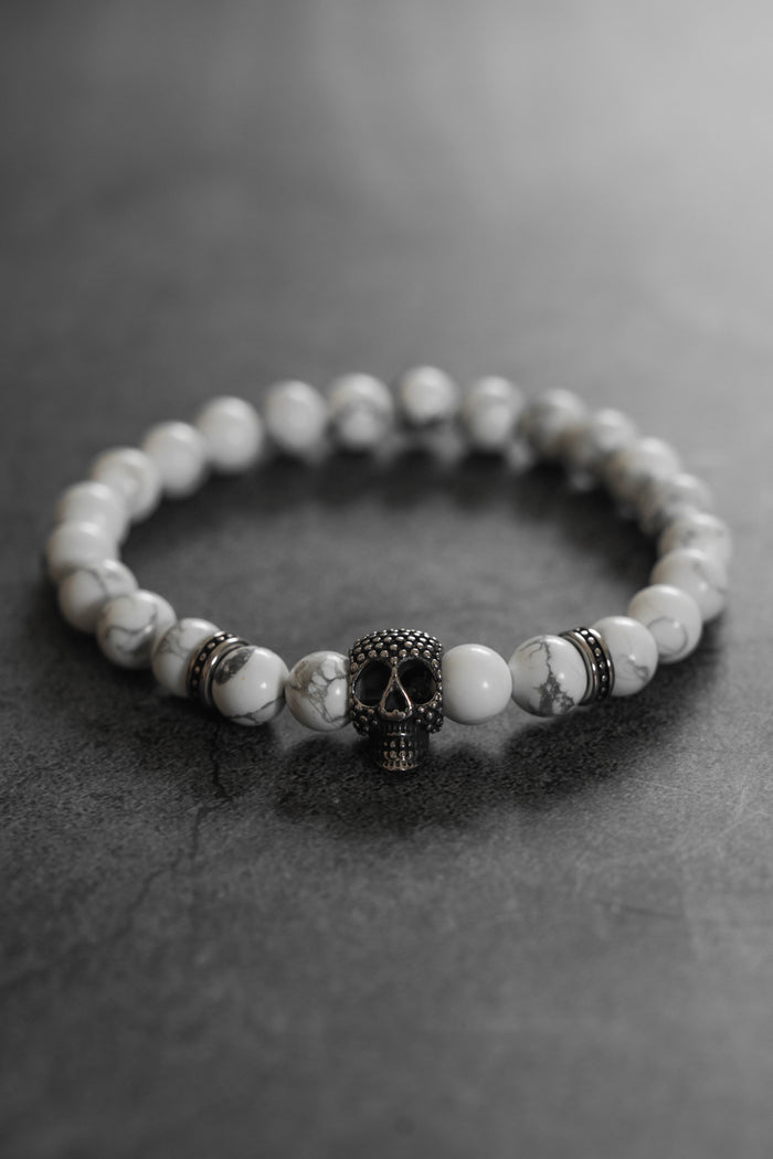 The Until Death Storm Cloud Bead Bracelet by Relentless Betrayal showcases white marbled beads and a bold metallic skull centerpiece, stylishly presented on a dark, textured surface.