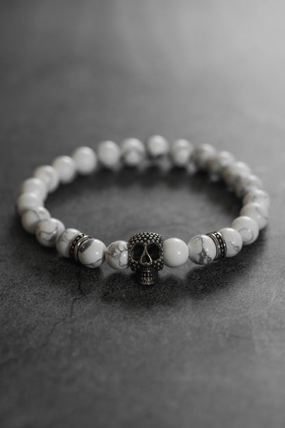 The Until Death Storm Cloud Bead Bracelet by Relentless Betrayal showcases white marbled beads and a bold metallic skull centerpiece, stylishly presented on a dark, textured surface.