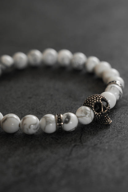 The Relentless Betrayal Until Death Storm Cloud Bead Bracelet, featuring white marbled beads and a metallic skull charm, is displayed on a dark, textured surface.