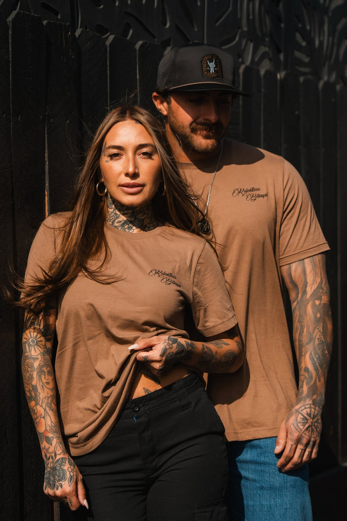A tattooed woman and man stand side by side wearing matching Vengeance Camel T-Shirts from Relentless Betrayal and dark pants. The man wears a cap, and they are set against a dark background. Sunlight casts empowering shadows on their faces, embodying resilience in their stance.