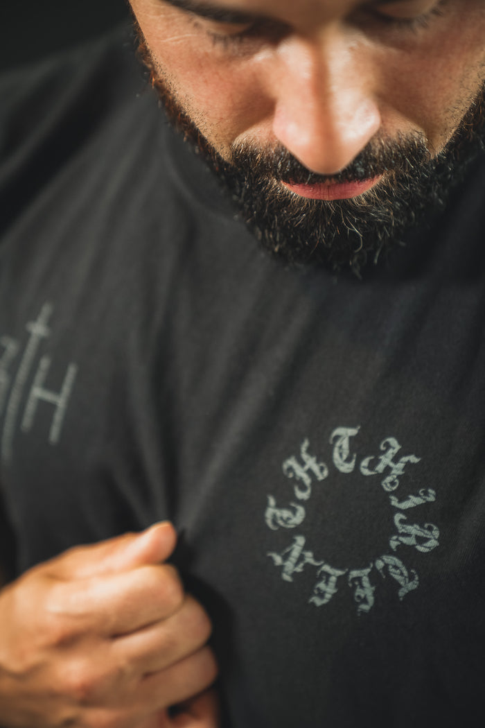 A bearded man in a black shirt looks down, holding the fabric near his chest. The 13th Victorious Dawn Premium Tee displays faint, stylized text in a circular pattern, symbolizing spiritual strength and a new dawn.