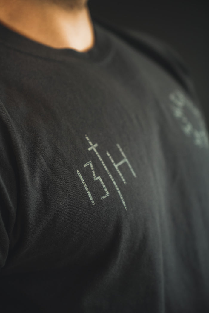 A close-up shows someone wearing the 13th Victorious Dawn Premium Tee, featuring white symbols and text on black fabric. The design stands out against a dark, blurred background, evoking spiritual strength.