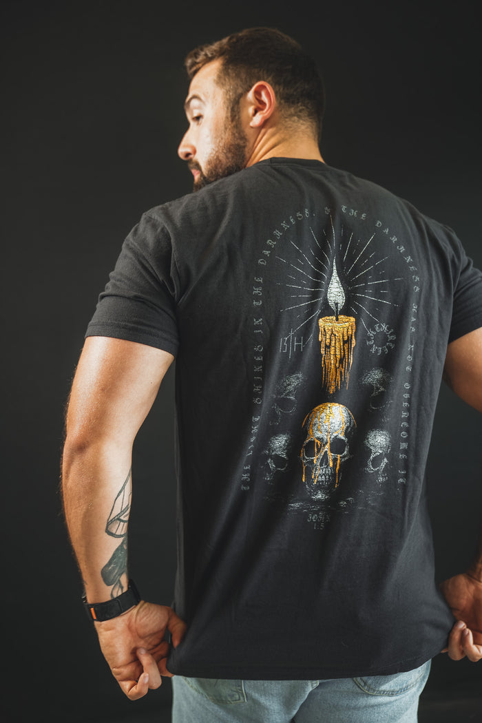 A bearded man stands sideways, displaying the back of his black 13th Victorious Dawn Premium Tee with artwork of a melting candle above an ornate skull, surrounded by text and small skulls, set against a dark background.