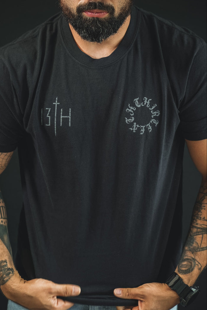 A bearded, tattooed man stands against a dark background, holding the hem of his Victorious Dawn Premium Tee by 13th, featuring bold faith-inspired graphics and exuding spiritual strength.