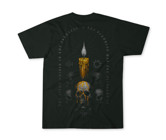 The 13th Victorious Dawn Premium Tee features a bold lit candle over a detailed skull, surrounded by faded skulls. Encircling text reads: The light shines in the darkness, & the darkness has not overcome it—symbolizing spiritual strength.