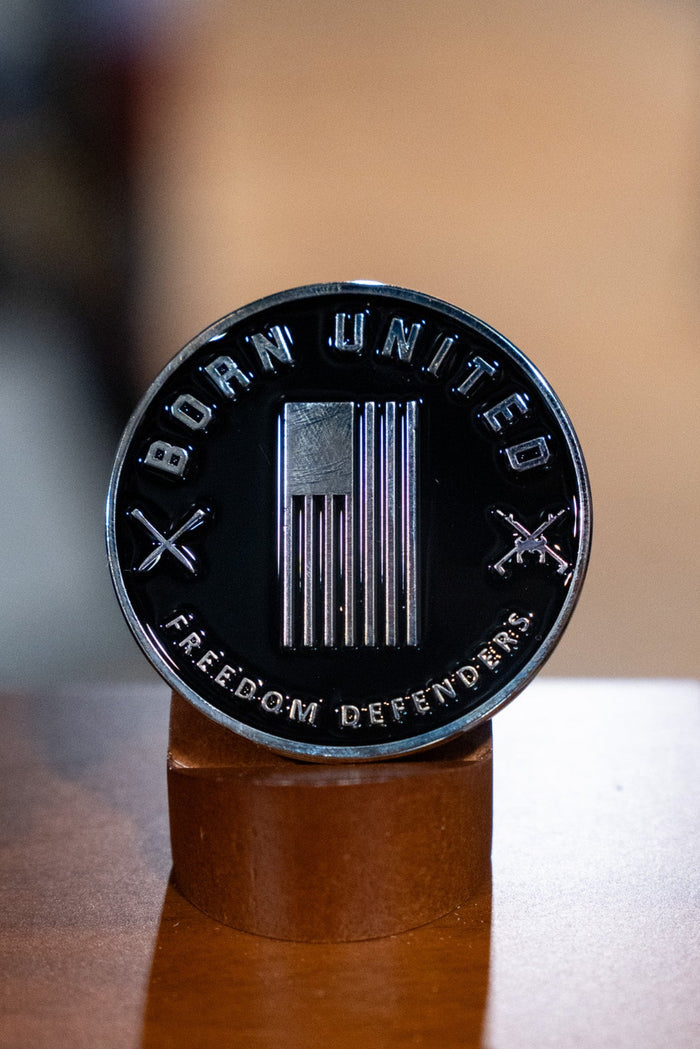 The Waking Lions Challenge Coin by Born United Private Label features an American flag, crossed rifles, and the words BORN UNITED and FREEDOM DEFENDER—a striking black collectible displayed on wood.
