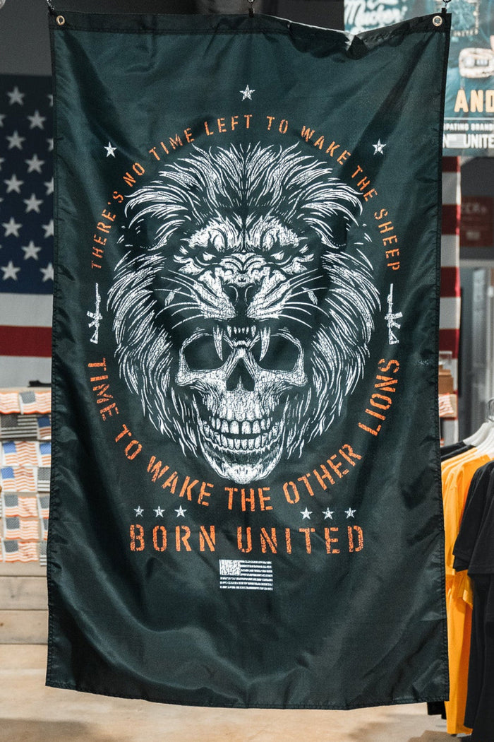 The Born United Private Label Waking Lions Flag features a black background with a roaring lion and skull, surrounded by the text: "There’s no time left to wake the sheep. Time to wake the other lions." T-shirts and American flags appear behind it.
