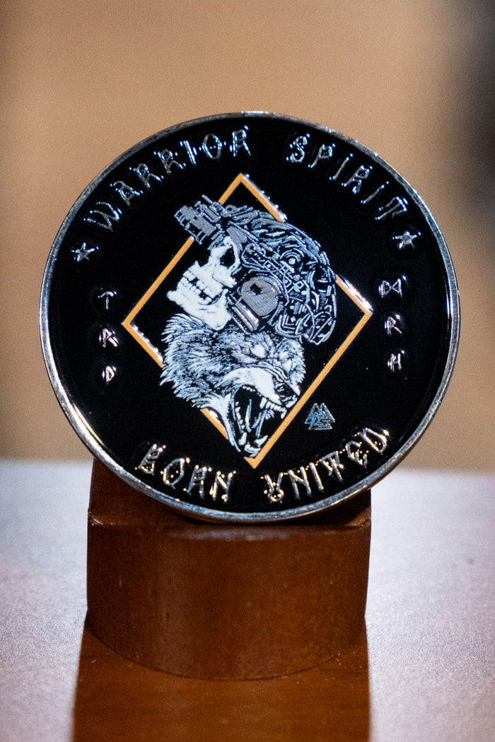 The Warrior Spirit Challenge Coin by Born United Private Label is a round black coin with a wolf’s head in a tactical helmet, "Warrior Spirit" inscribed at the top, and "BORN UNITED" at the bottom, displayed upright on a wooden stand.