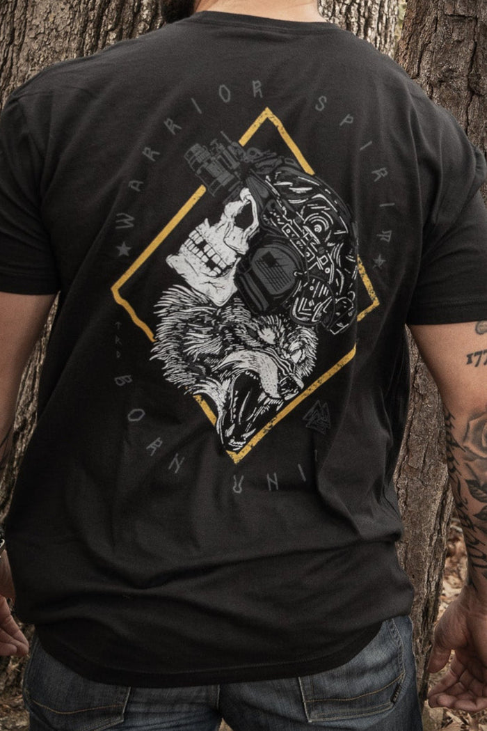 A man stands in a forest with his back to the camera, wearing Born United Private Labels Warrior Spirit black t-shirt, featuring a wolf graphic and text. His tattooed left arm complements the strength of character amidst tall trees and fallen leaves.