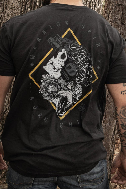 A man stands in a forest with his back to the camera, wearing Born United Private Labels Warrior Spirit black t-shirt, featuring a wolf graphic and text. His tattooed left arm complements the strength of character amidst tall trees and fallen leaves.