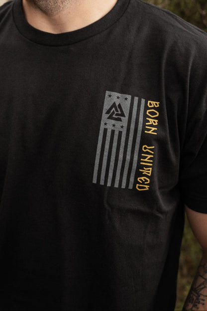 In a wooded area, a bearded man wearing a black Warrior Spirit T-shirt by Born United Private Label stands, evoking the strength of First Responders. He looks to the side, surrounded by trees and fallen leaves that symbolize resilience.