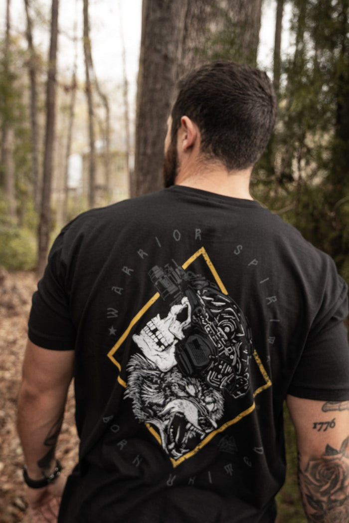 A tattooed person walks in a forest, wearing a Born United Private Labels Warrior Spirit black T-shirt. The back features a skull and wolf graphic, symbolizing the strength of first responders with the words Warrior Spirit.